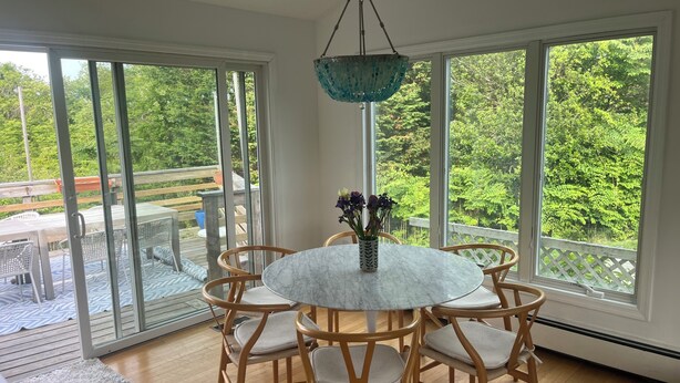 A dining area with a round table, chairs, a hanging light fixture, and large windows offering a view of greenery.