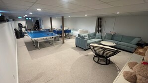 A room with a ping pong table, a sofa, a round table, and a teddy bear.