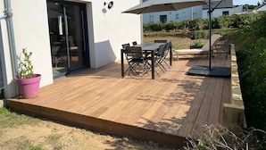Una terraza de madera con una mesa, sillas, una planta en maceta y un ventanal que da a un espacio interior.