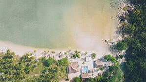 Vue aérienne d’une propriété située en bord de mer, avec une piscine, plusieurs bâtiments de style bungalow et une rangée dense de palmiers.
