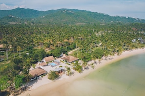 Eine Vogelperspektive zeigt einen Küstenort mit strohgedeckten Bungalows, einem Sandstrand und im Hintergrund dichte Palmen und Hügel.