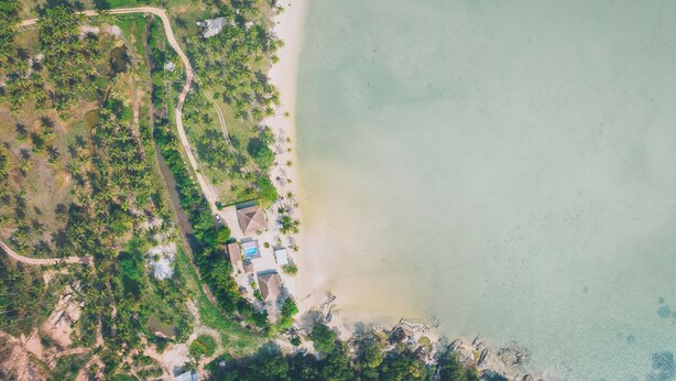 Vue aérienne d’une propriété située sur la côte, avec une plage, des palmiers et un petit ensemble de bâtiments dotés d’une piscine.