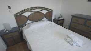 Een net opgemaakt bed met een witte deken, een houten hoofdbord met een geweven patroon, een nachtkastje met een lamp en een ladekast.