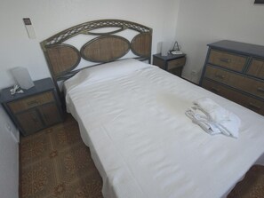 Ein ordentlich bezogenes Bett mit weißer Decke, ein Holz-Kopfteil mit gewebtem Muster, ein Nachttisch mit Lampe und eine Kommode mit Schubladen.