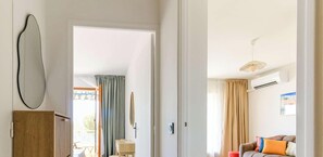 A room with a wooden cabinet, a small table, a sofa with cushions, a hanging light fixture, and a view through an open door. - Charming apartment, sea view - 4 guests (Bormes-les-Mimosas)
