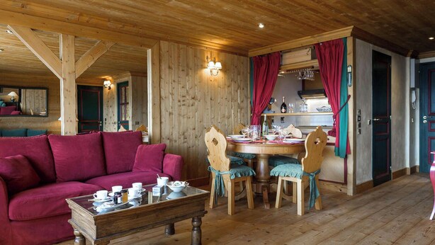 A cozy room with a wooden ceiling, a red sofa, a wooden dining table with chairs, and a kitchen area visible through a doorway. - Ski-in/ski-out apartment, view - 4 guests (Courchevel)