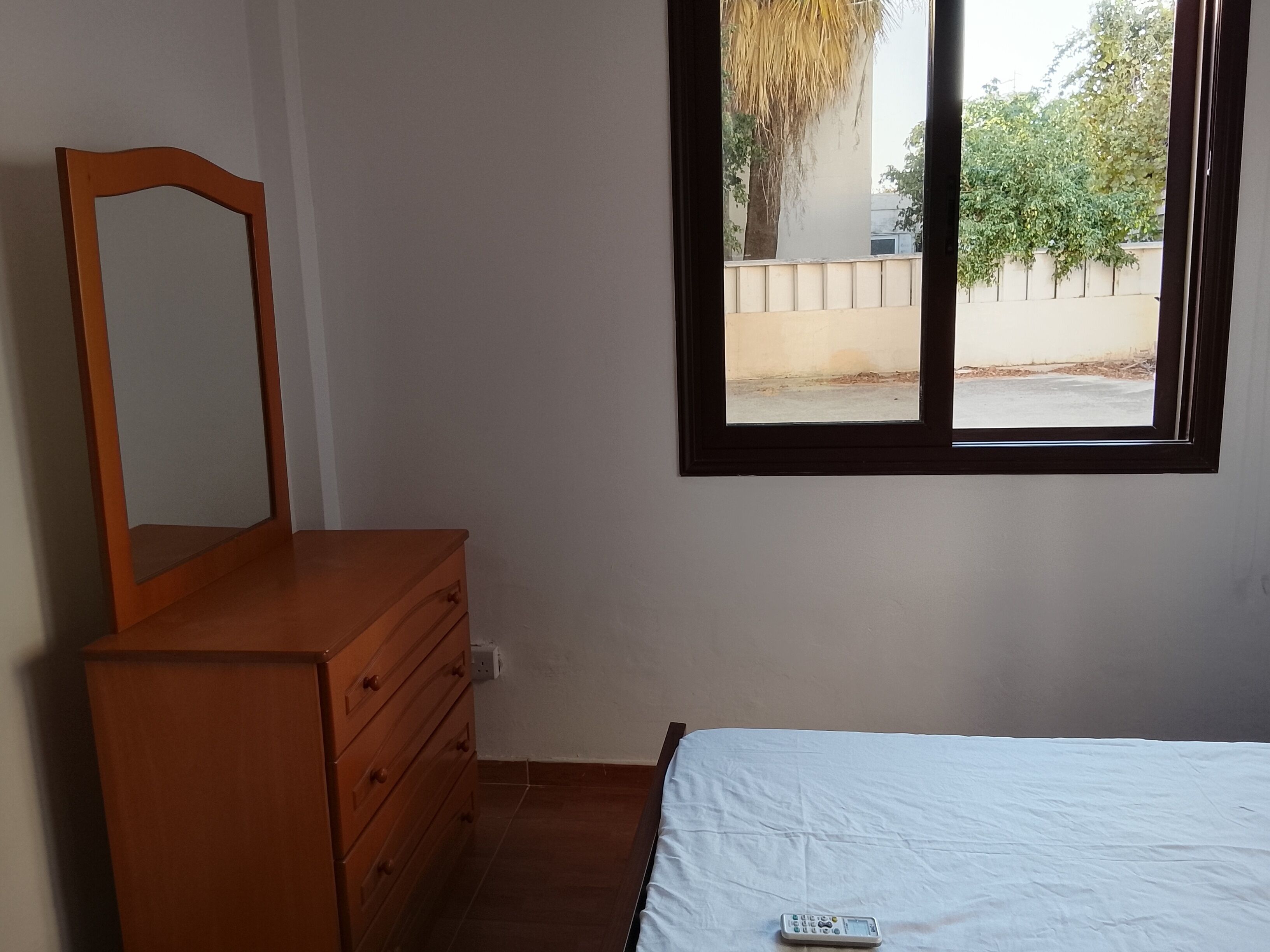Un dormitorio con un aparador de madera, una cama con sábana blanca y una ventana que ofrece vistas a la calle y árboles.