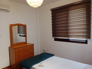 Ein Schlafzimmer mit einem Holzkoffer, einem Bett mit blauer Decke, einer Deckenleuchte und einem Fenster mit Jalousien.