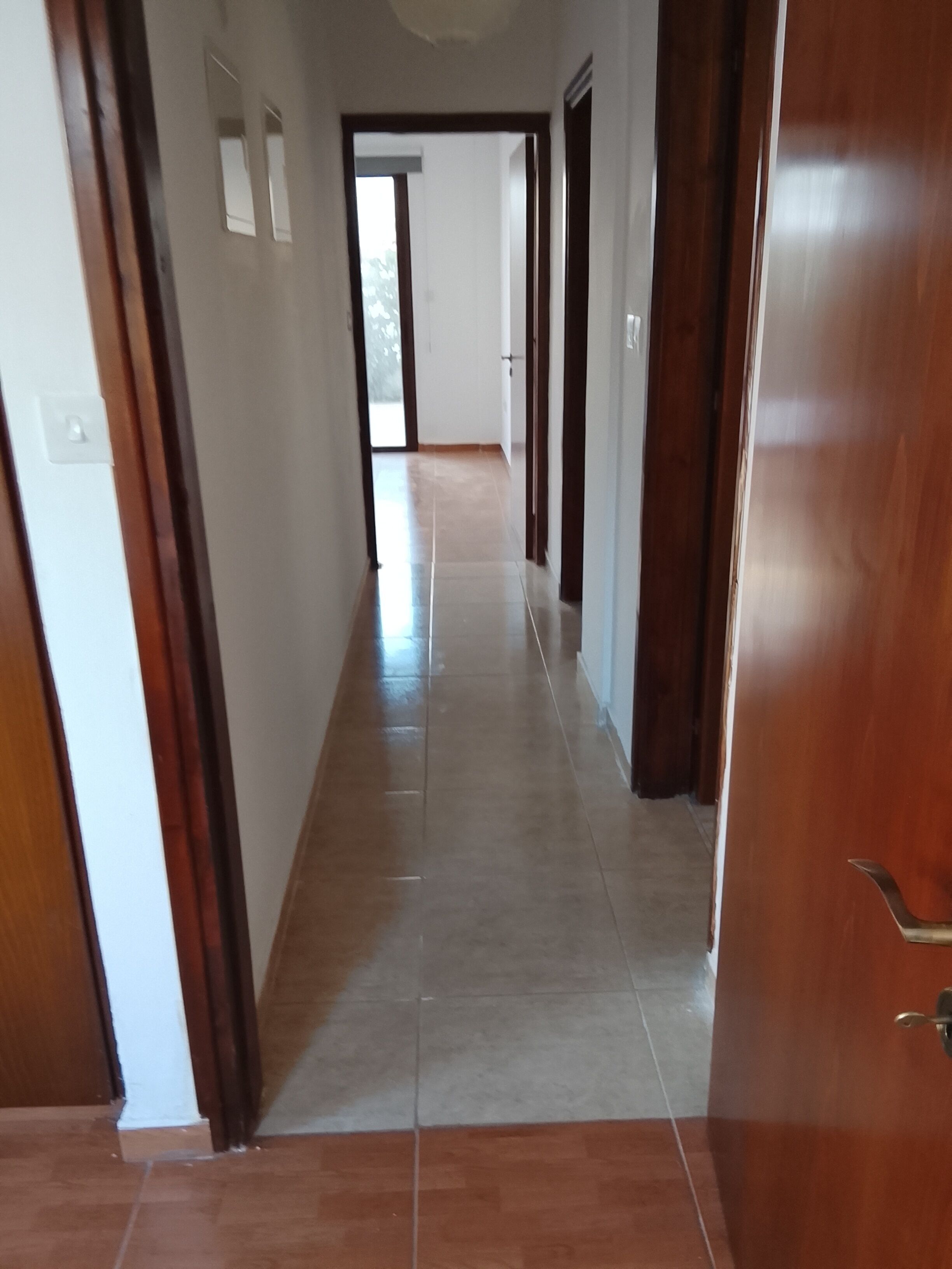 A hallway with a wooden door on the right, a white wall with a light switch, and a tiled floor.