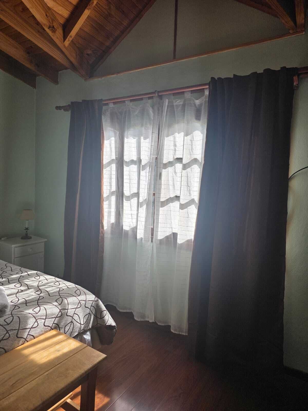A bedroom with a bed, a nightstand, a wooden bench, and a window with curtains.