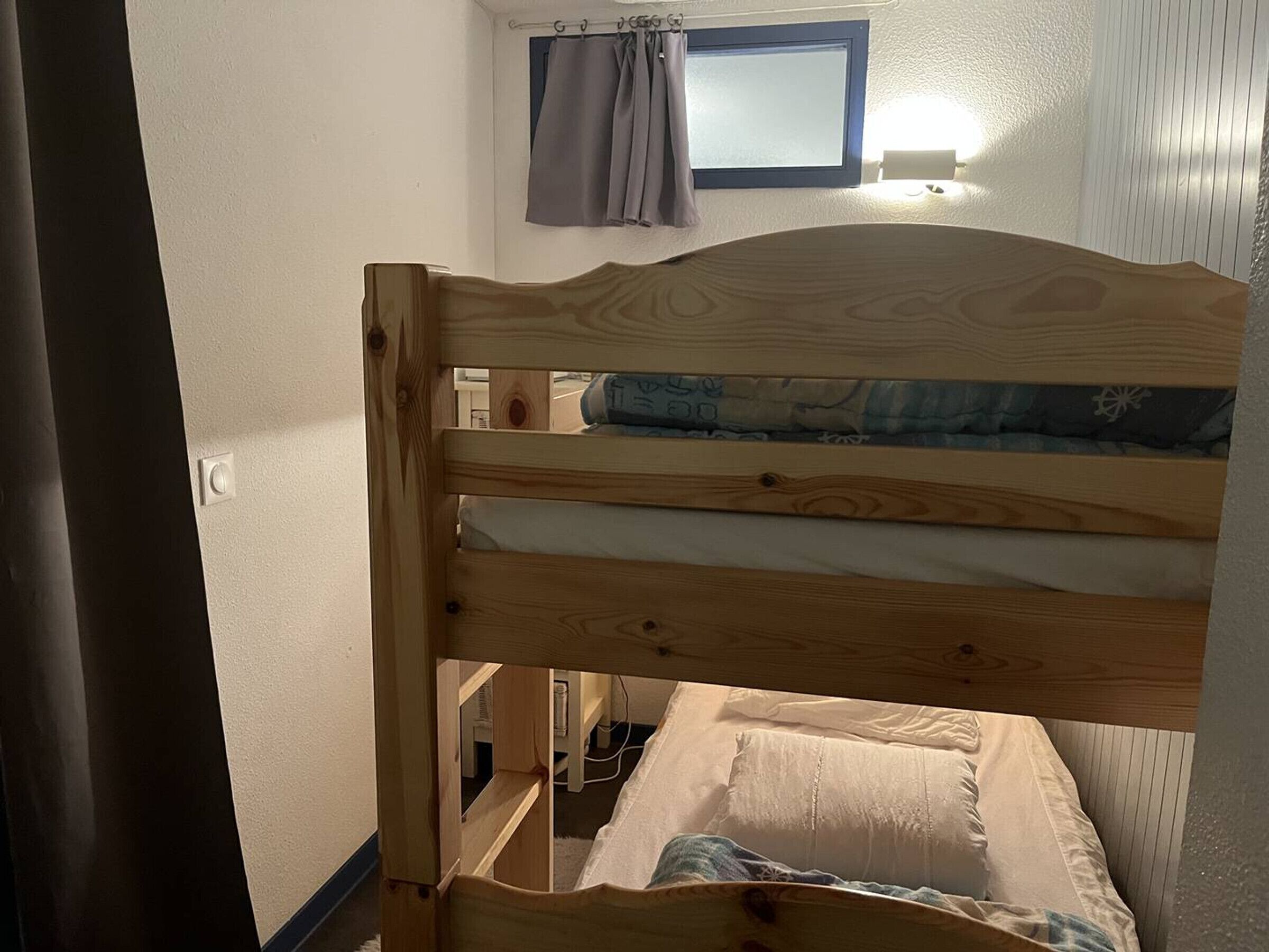 A bunk bed with a wooden headboard, a window with curtains, and a wall-mounted light fixture.