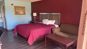 A hotel room with a bed, a sofa, a small table, a nightstand with a lamp, and a painting on the wall.