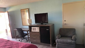 A room with a bed, a desk with a computer, a television, a microwave, and a patterned armchair.