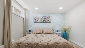 A bedroom with a large bed, a nightstand, a vase with flowers, and a wall-mounted painting.