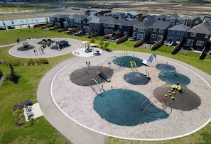 A playground with various play structures surrounded by a gravel area, set against a backdrop of residential buildings. - Pristine Suite Near Fish Creek Park & Horse Events (500 Mbps WiFi + Workspace) (Calgary)