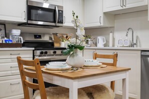 A modern kitchen with white cabinetry, a stainless steel microwave, a stove, and a dining table set with plates and a vase of flowers.