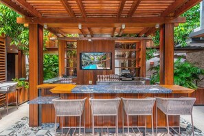 Eine Bar mit Holztresen, einem Fernseher, Barhocker und Blick auf den Pool.