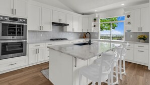 Een moderne keuken met witte kasten, roestvrijstalen apparatuur, een centraal eiland met barstoelen en een groot raam met uitzicht.