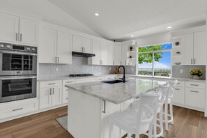 A modern kitchen with white cabinetry, stainless steel appliances, a central island with bar stools, and a large window offering a view. - Draper Skyline Retreat | Hot Tub & 6BDR (Draper)