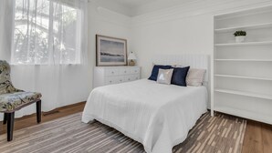 Een slaapkamer met een bed, een stoel, een dressoir, een raam met dunne gordijnen, een ingelijst kunstwerk en een witte plank.