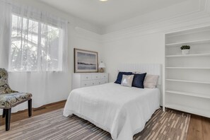 A bedroom with a bed, a chair, a dresser, a window with sheer curtains, a framed picture, and a white shelving unit. - Draper Skyline Retreat | Hot Tub & 6BDR (Draper)