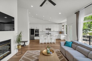 A modern living room with a fireplace, a sofa, a coffee table, and a view of the kitchen and dining area. - Draper Skyline Retreat | Hot Tub & 6BDR (Draper)