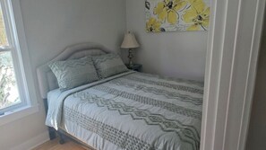 Un dormitorio con cama, almohadas, una mesita de noche, lámpara y una obra de arte floral en la pared.