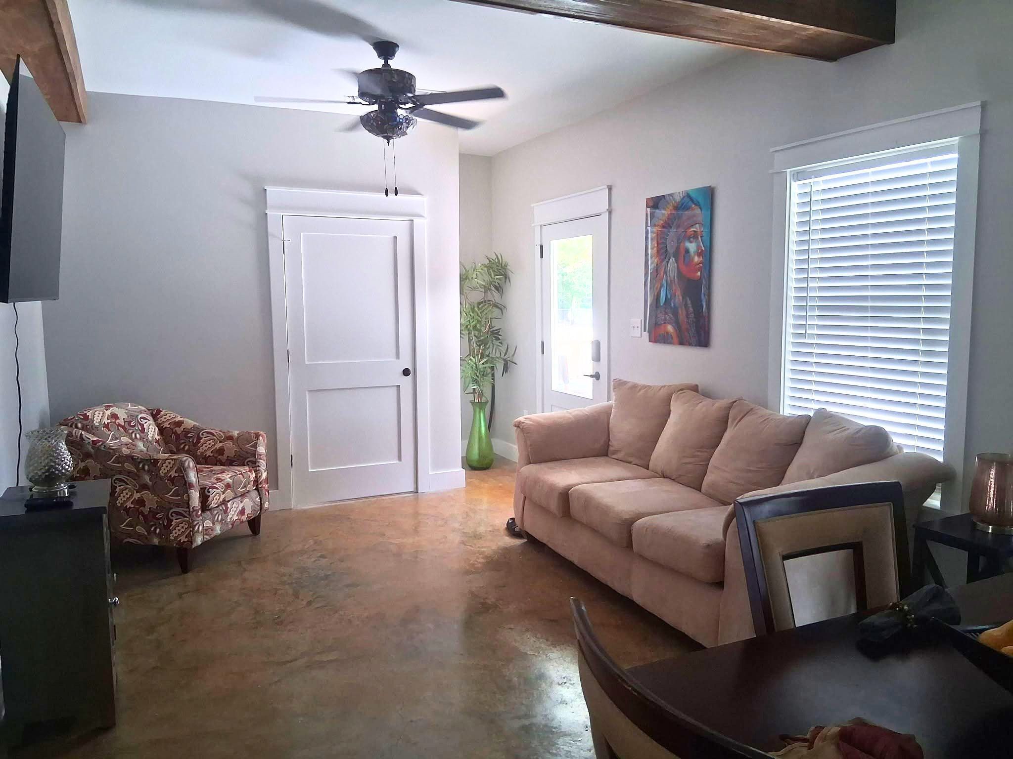 A living room with a beige sofa, patterned armchair, and a dining table. There is a ceiling fan, a green plant, and a painting on the wall.