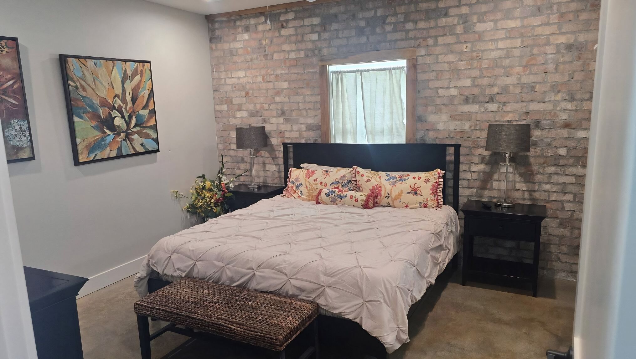A bedroom with a brick wall, a bed with a floral bedspread, two nightstands, and a window with curtains.