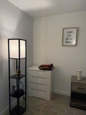 Um quarto com uma luminária de chão alta, uma cômoda branca, uma pequena mesa com uma xícara e um pôster motivacional na parede.