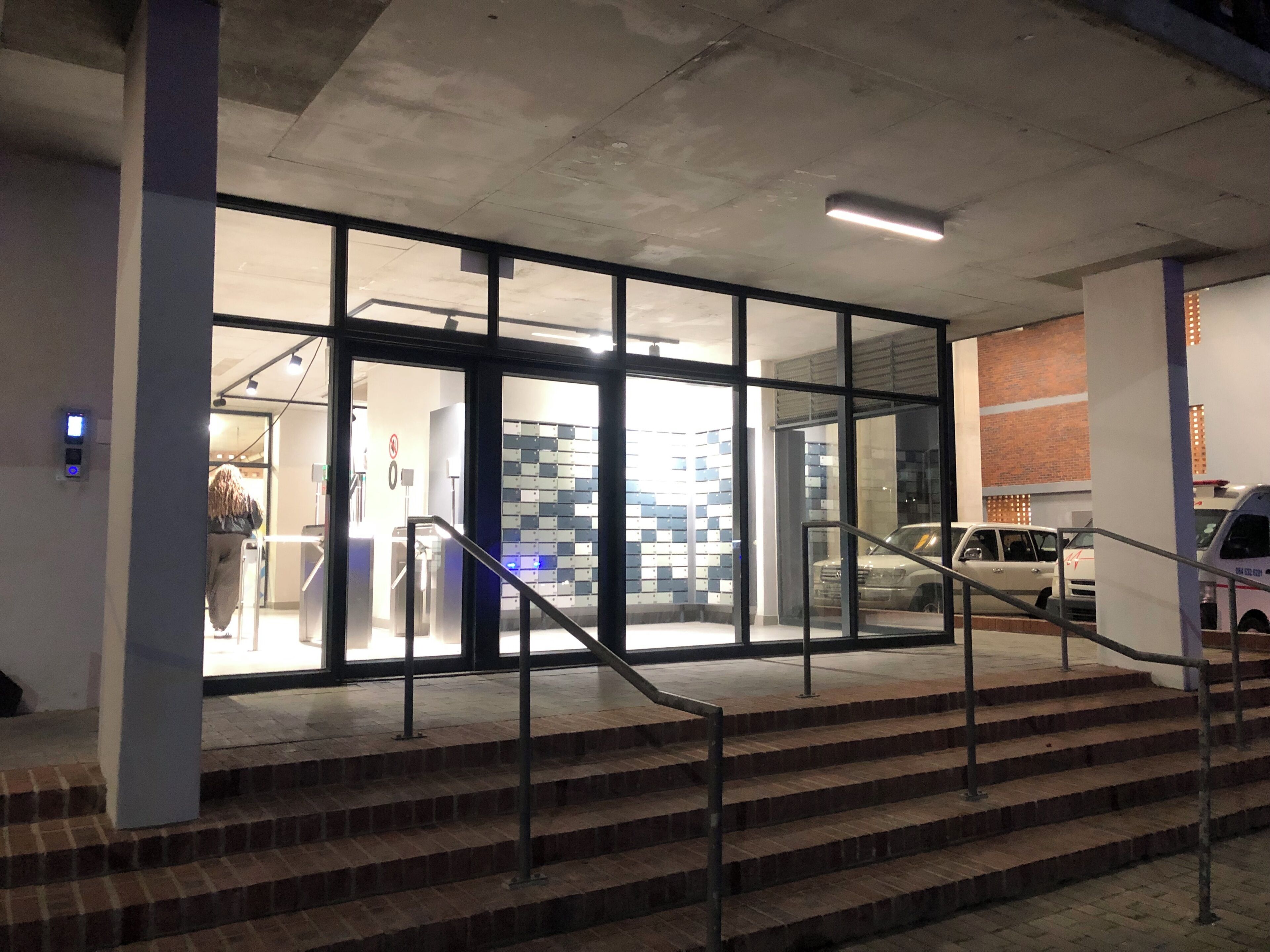 Une entrée d’immeuble avec des portes vitrées, un escalier et un espace de stationnement avec des voitures.