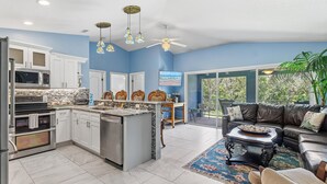 A modern kitchen with white cabinets, a central island, and a dining area with a black leather sofa and a coffee table. - The Flying Mermaid (Edgewater)