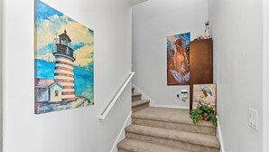 A staircase with beige carpet, white walls, and two paintings: one of a lighthouse and another of a woman. - The Flying Mermaid (Edgewater)