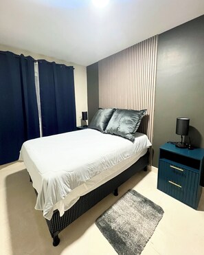 A bedroom with a bed, a nightstand, a lamp, a rug, and a wall with vertical blinds.