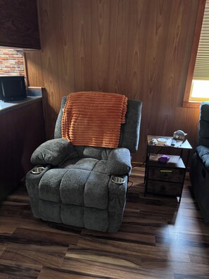 Ein gemütliches Zimmer mit einem gepolsterten Sessel, einem kleinen Holztisch und einem Fenster mit Jalousien.