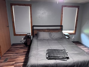 Ein Schlafzimmer mit einem Bett, zwei Fenstern mit Jalousien, einem Nachttisch und einem Beistelltisch.