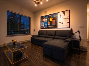 Ein modernes Wohnzimmer mit einer Eckcouch, einem Glastisch, einem Schreibtisch mit Lampe und einem großen Fenster mit Jalousien.