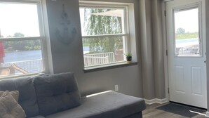 Una habitación con un sofá gris, una ventana con vista a un lago y una puerta blanca.