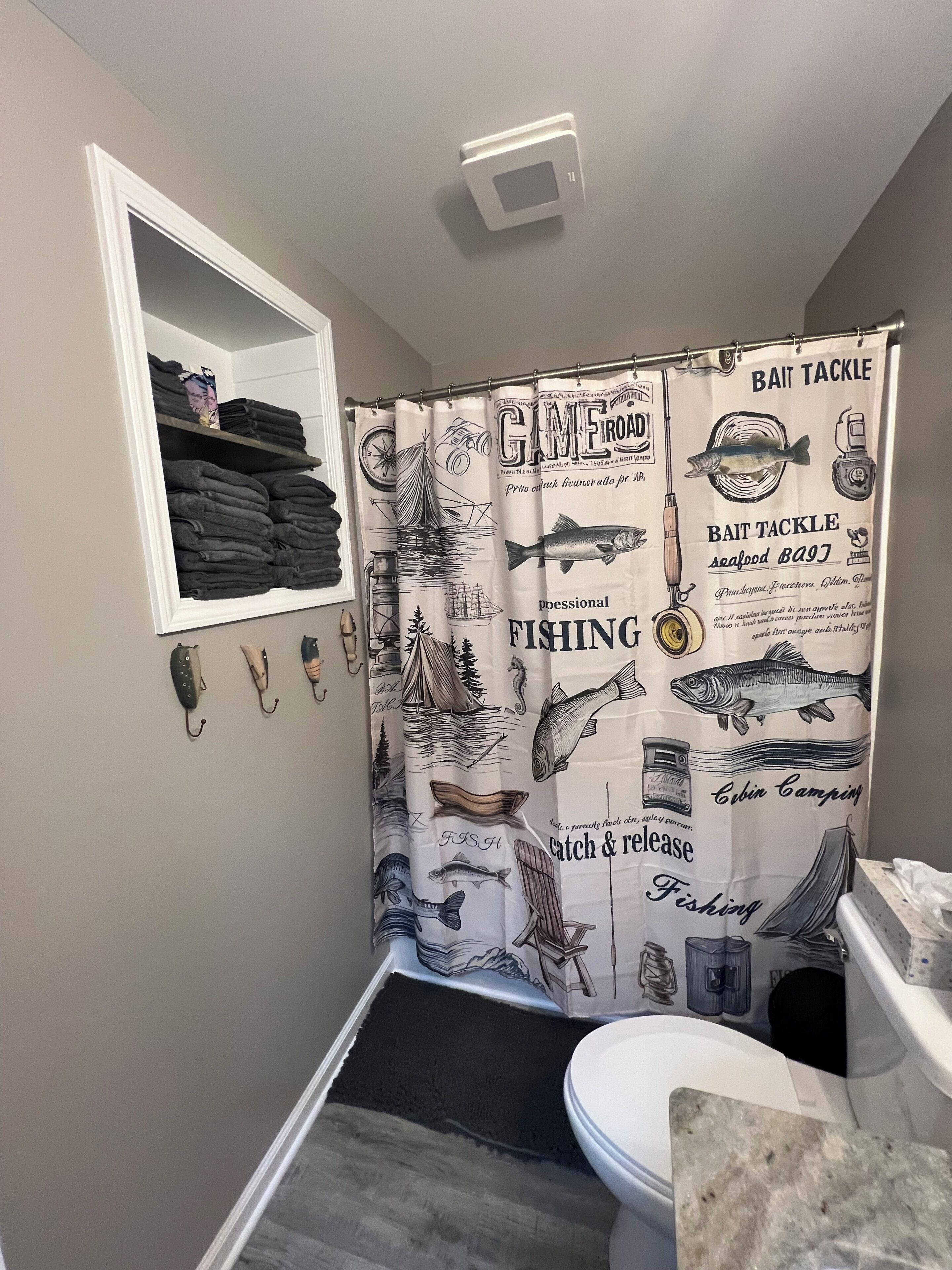 A bathroom with a fishing-themed shower curtain, a shelf with folded towels, and a toilet with a marble countertop.