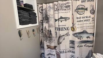 A bathroom with a fishing-themed shower curtain, a shelf with folded towels, and a toilet with a marble countertop.