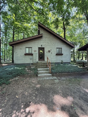 A single-story cabin with a front porch, a welcome sign, and a small window on each side.