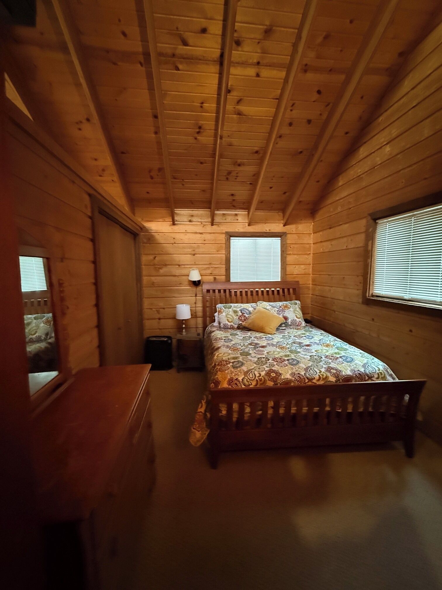 A wooden cabin bedroom with a bed, bedside lamp, and a window with blinds.