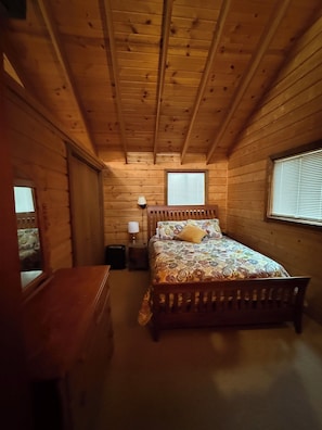 A wooden cabin bedroom with a bed, bedside lamp, and a window with blinds.