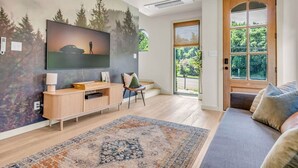 Un salon moderne doté d’un téléviseur à écran plat, d’un meuble de rangement en bois, d’un canapé et d’une grande fenêtre donnant sur un paysage verdoyant.