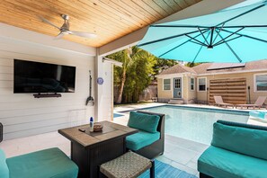 Uma área externa coberta com piscina, móveis de vime, ventilador de teto e televisão na parede.