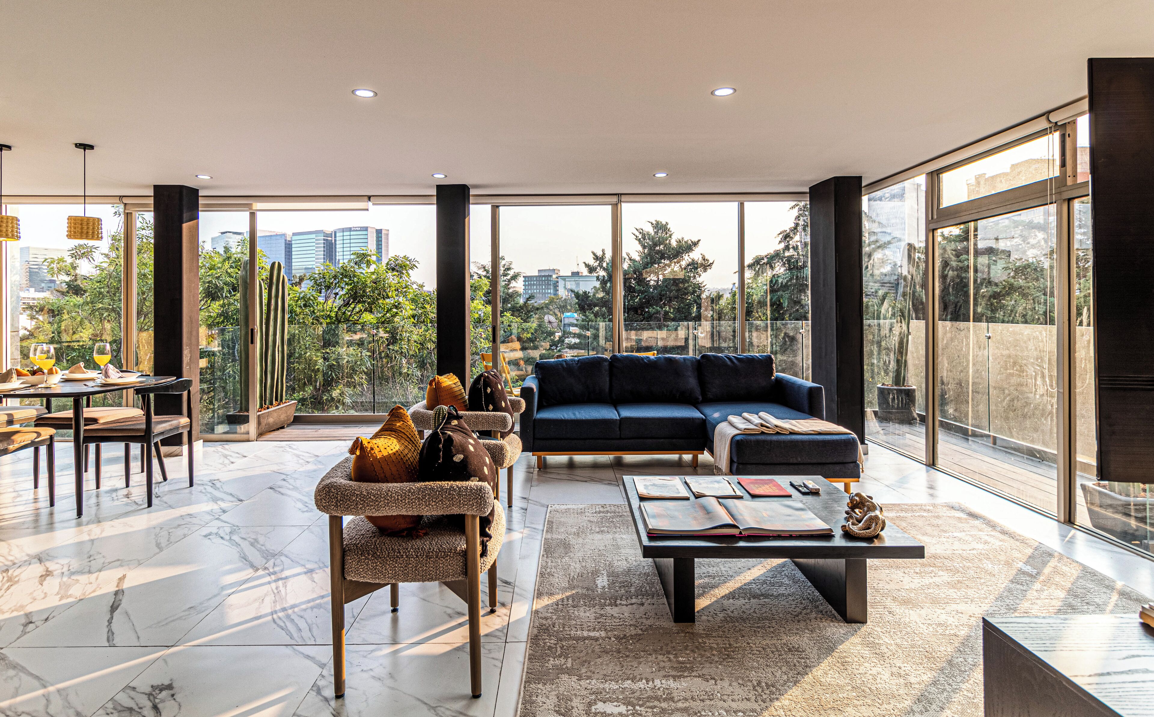 A spacious living area with a sofa, coffee table, and dining table, featuring large windows and a city view.