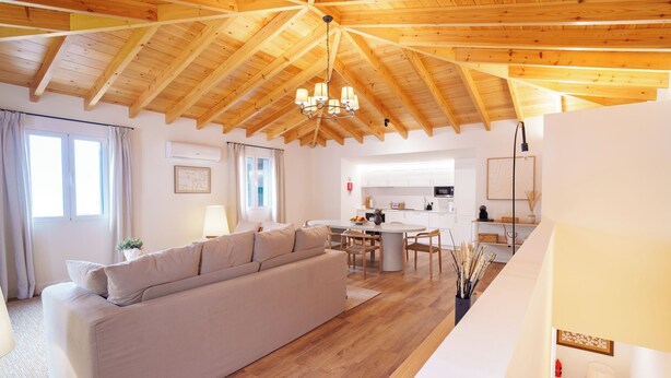Amplia sala de estar con techo de madera alto, un sofá, una mesa de comedor y una cocina al fondo.