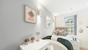 Et moderne soveværelse med seng, natbord, lampe og et vindue med udsigt til bygninger.