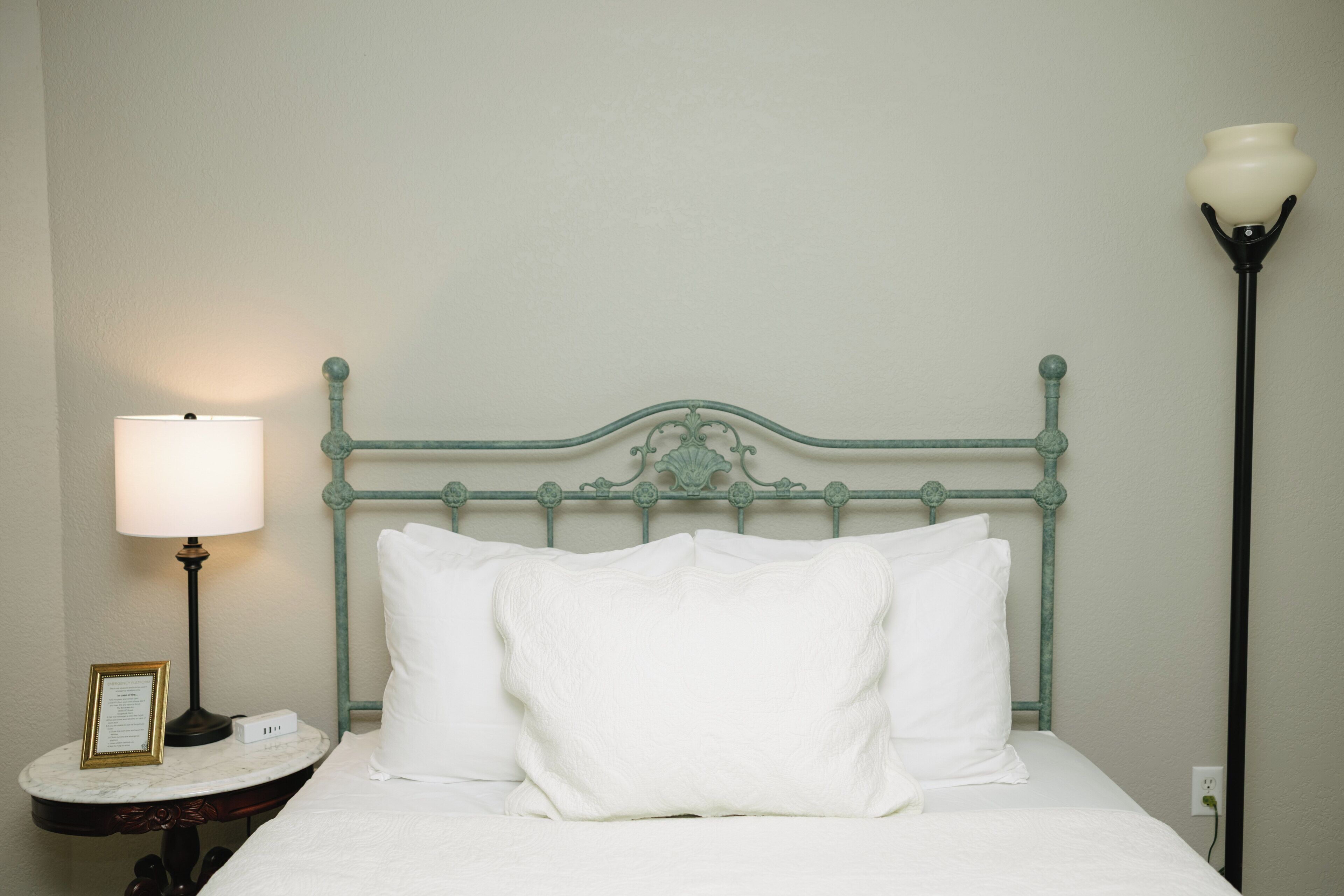 Un lit bien fait, avec du linge de lit blanc, une tête de lit en métal, une table de chevet avec une lampe et une prise électrique au mur.