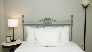 Un lit bien fait, avec du linge de lit blanc, une tête de lit en métal, une table de chevet avec une lampe et une prise électrique au mur.